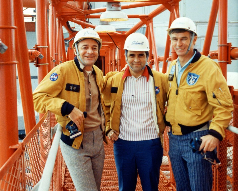 The three Apollo 17 astronauts in yellow jackets and white hard hats, standing on an orange steel gantry walkway at the launch pad