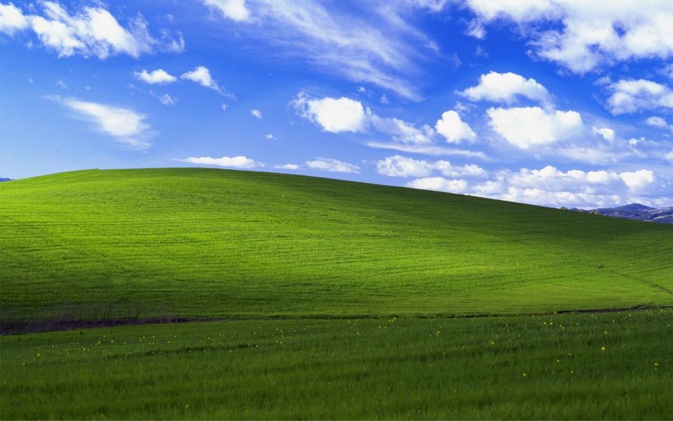 A smooth green hill rolling under a deep blue sky with scattered white clouds – the ‘Bliss’ desktop wallpaper.