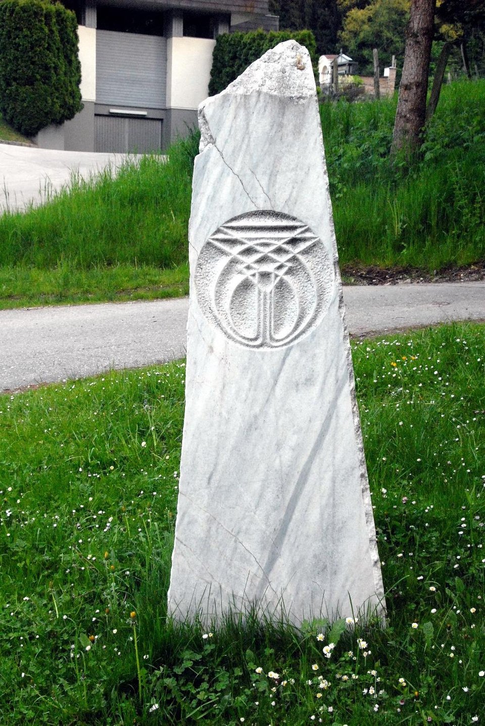 A tall white marble standing stone with a carved interlaced cosmogram, set in grass beside a road.